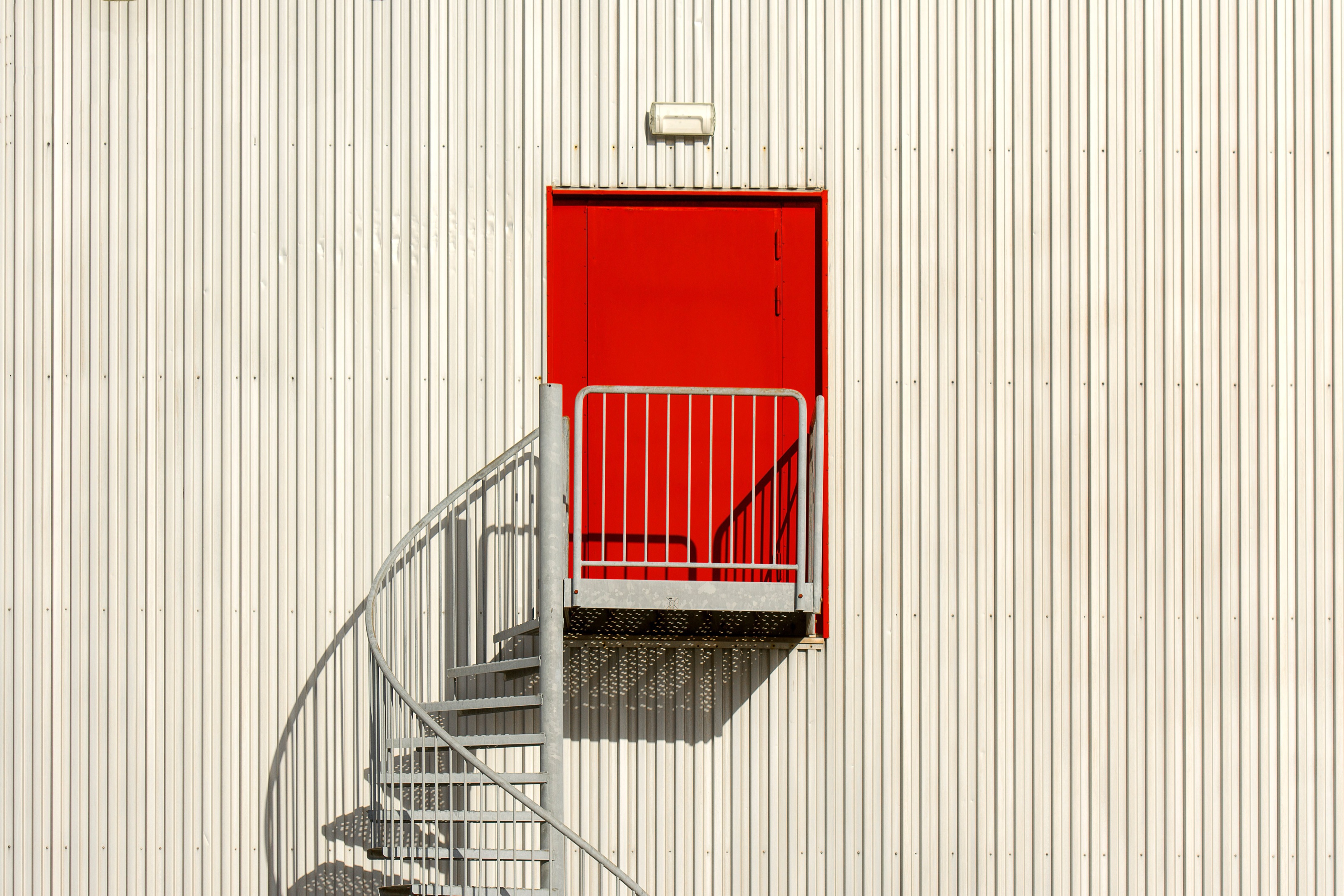 A red door and a spiral staircase on a white corrugated facade